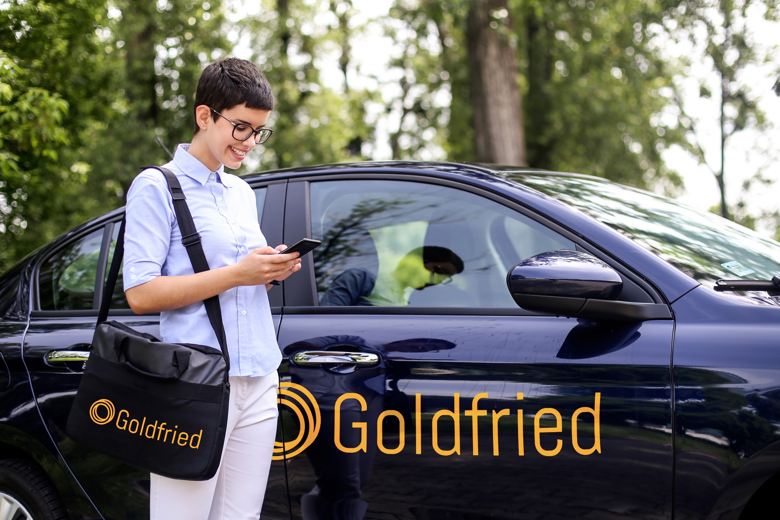 Mitarbeiterin von Goldfried mit Umhängetasche und Smartphone vor einem dunkelblauen Firmenwagen mit Goldfried Logo